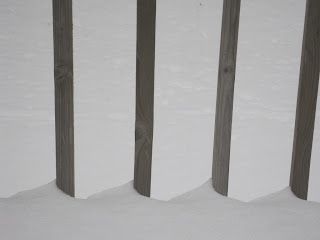 Snow accumulating between wooden fence posts.